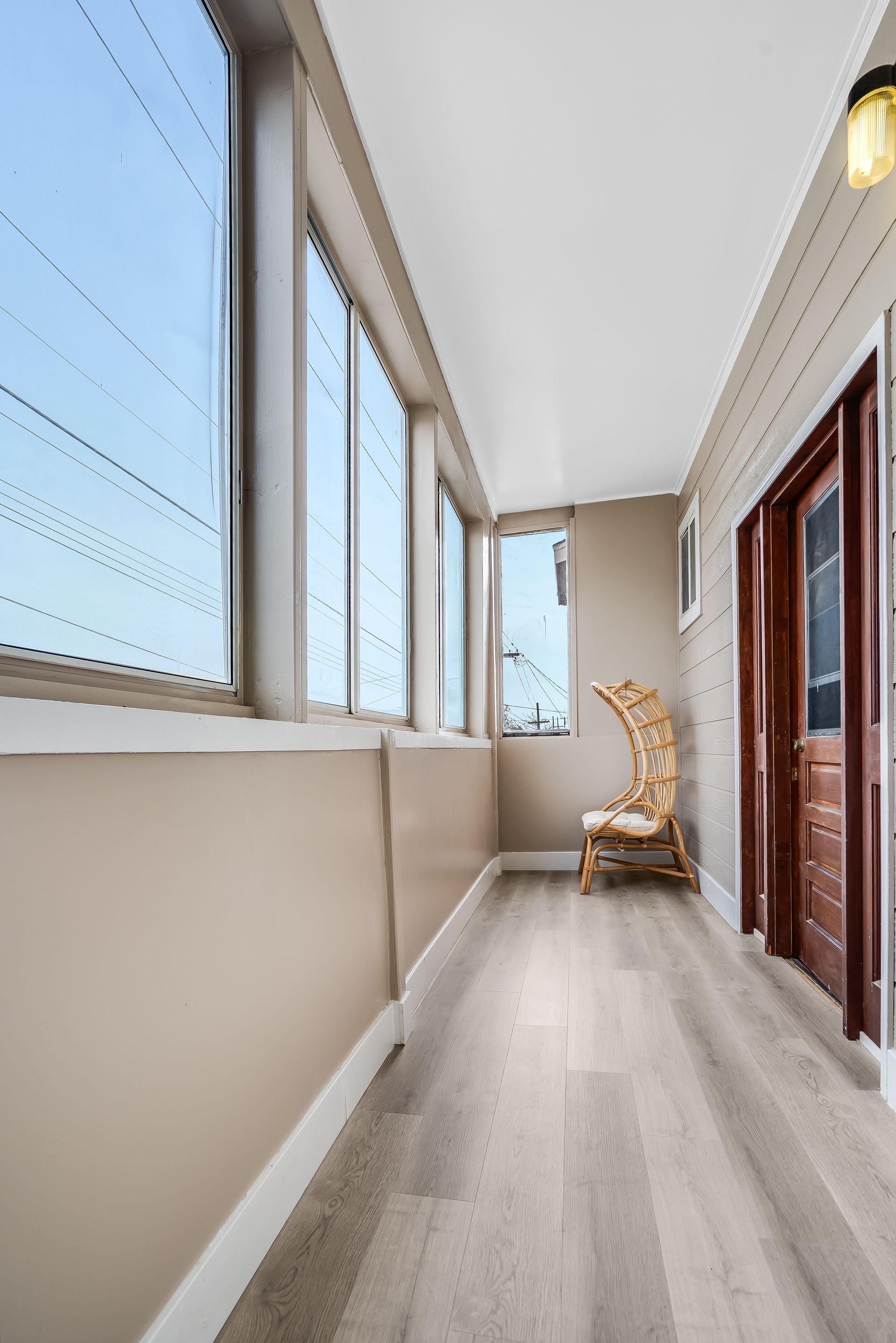 Long, bright hallway with large windows and a wicker chair. Light wood floors and a wooden door.