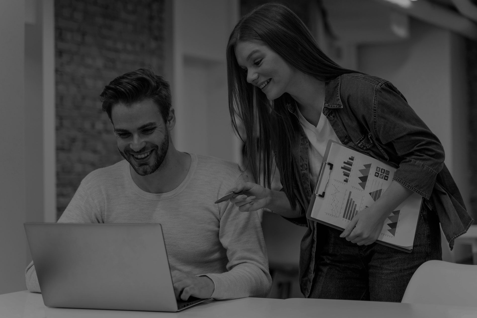 Two coworkers smiling at a laptop, one standing with a notebook in a bright office.