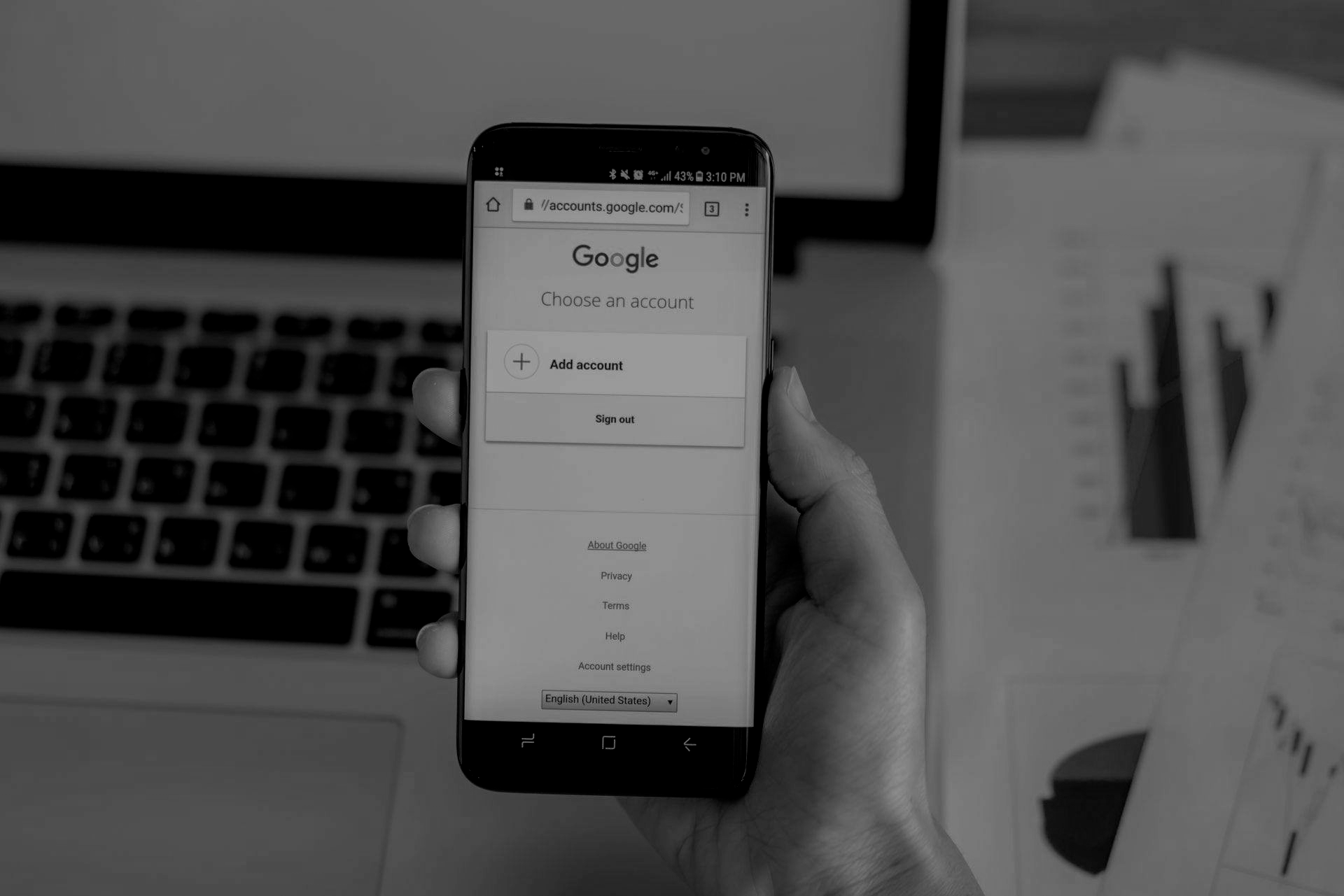 Person holding a smartphone, displaying the Google account login page. Laptop and paperwork in background.