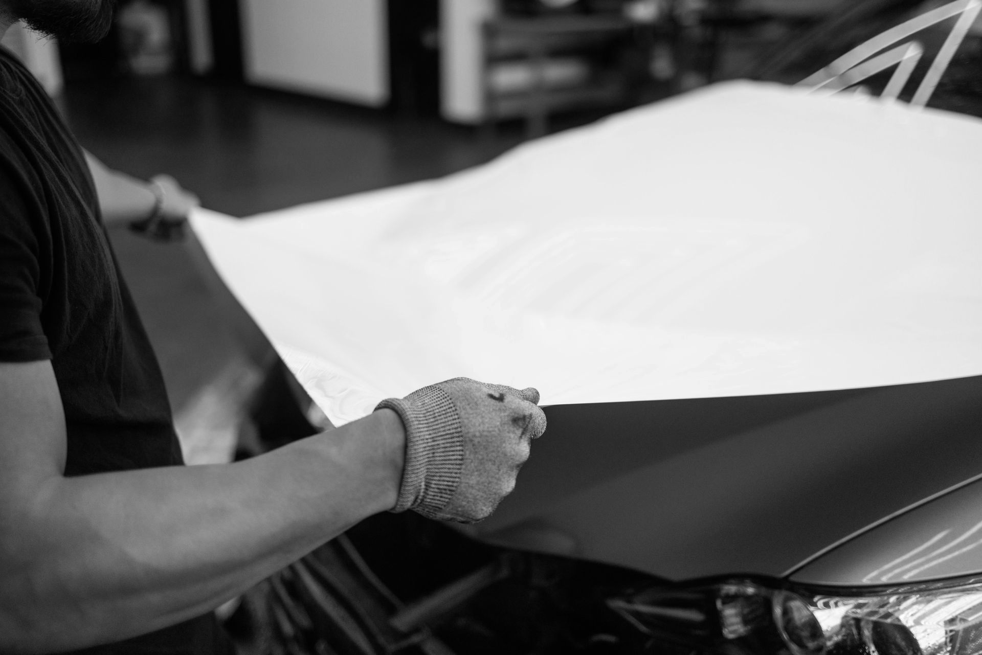 Person applying white vinyl wrap to a car hood. Gloves, interior garage.