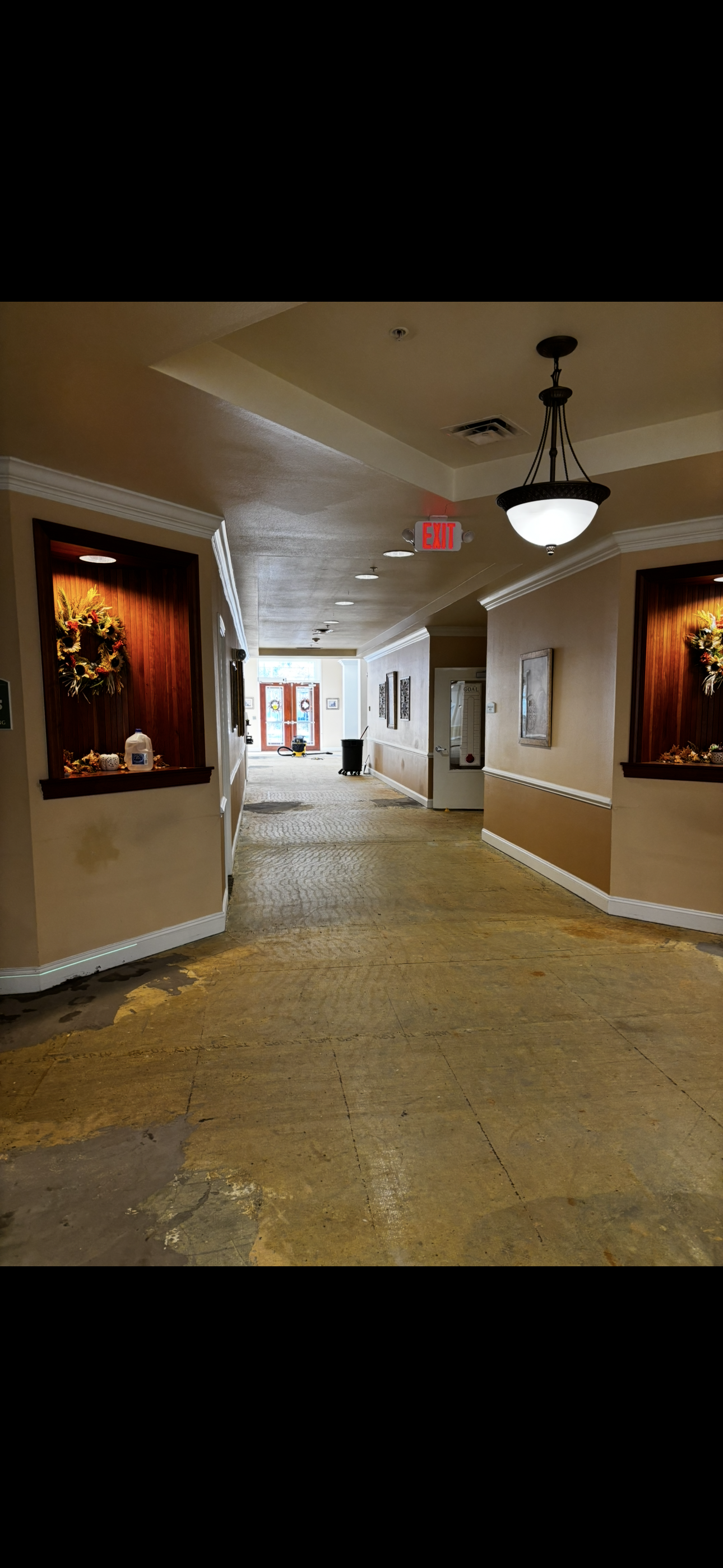 Hallway with stained floor and damaged walls. Overhead lighting, two display alcoves. Exit sign visible.