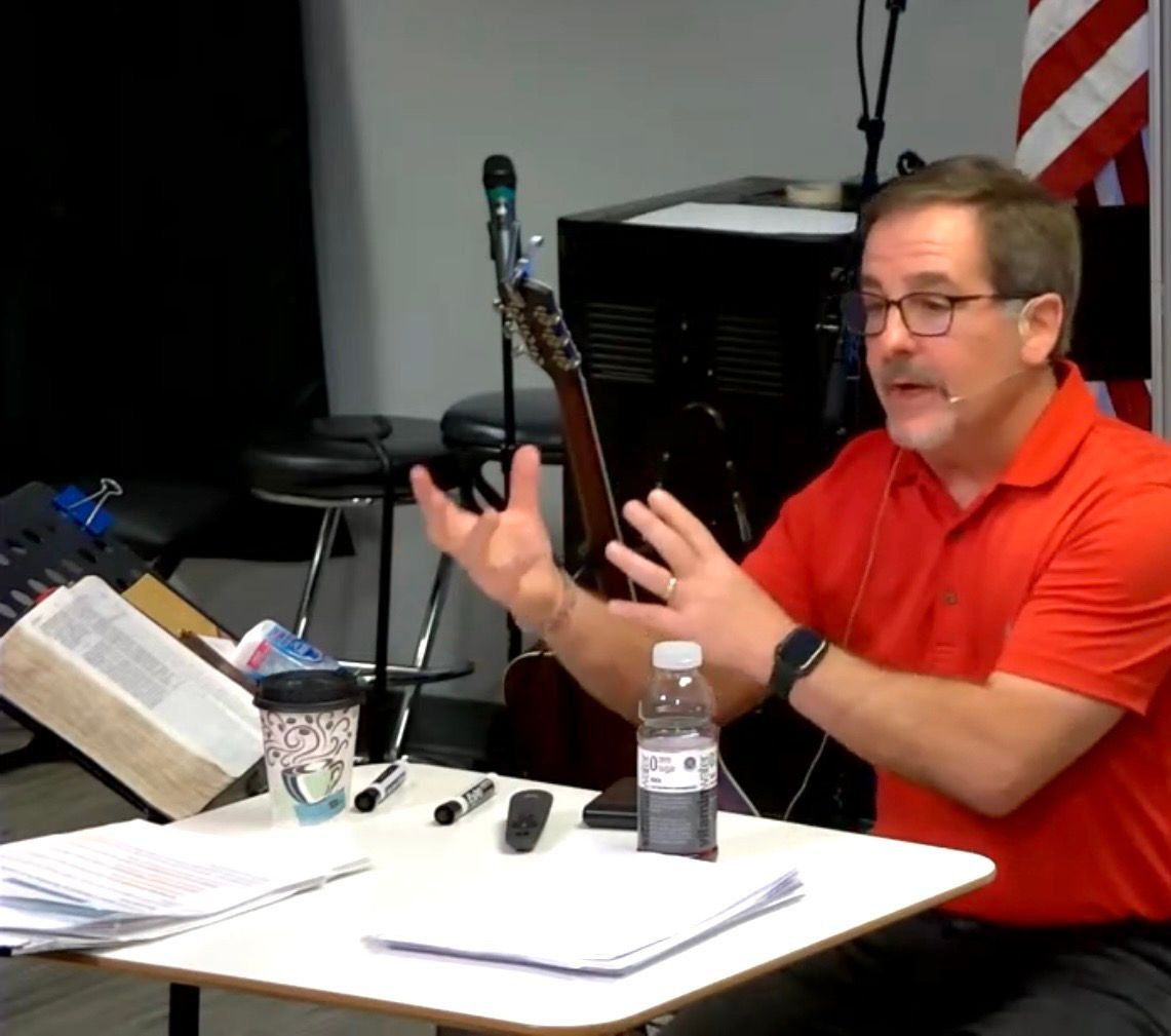 Man in red polo speaking at a table with papers, microphone, and water bottle in a classroom setting
