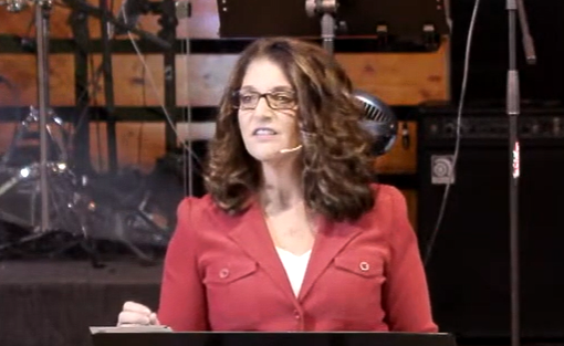 Woman speaking into a microphone on a stage, wearing glasses and a white lace top.
