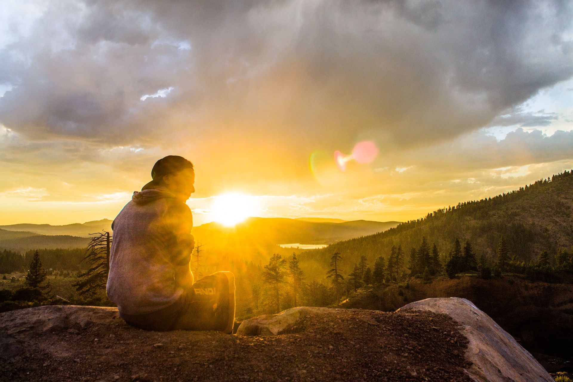 Person sitting on a rock at sunset, overlooking a golden mountain valley under cloudy skies