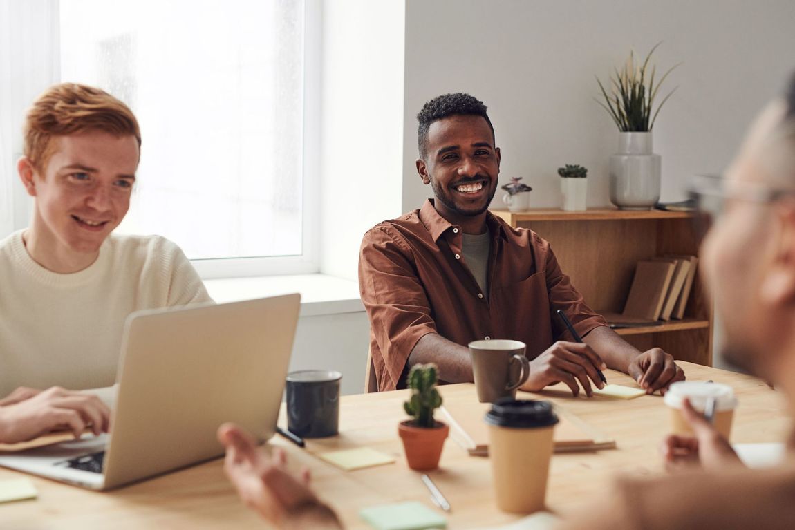 People seated around a table in a bright meeting room, smiling and using laptops and coffee cups.