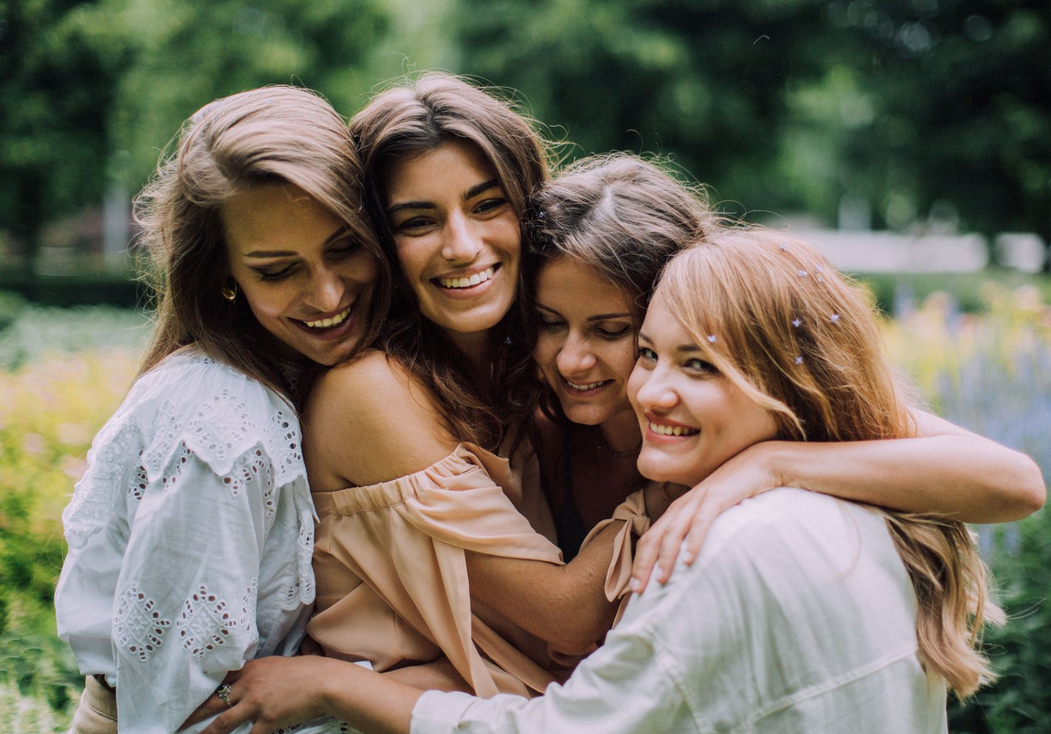 Four friends hugging and smiling in a green park, wearing light-colored tops.