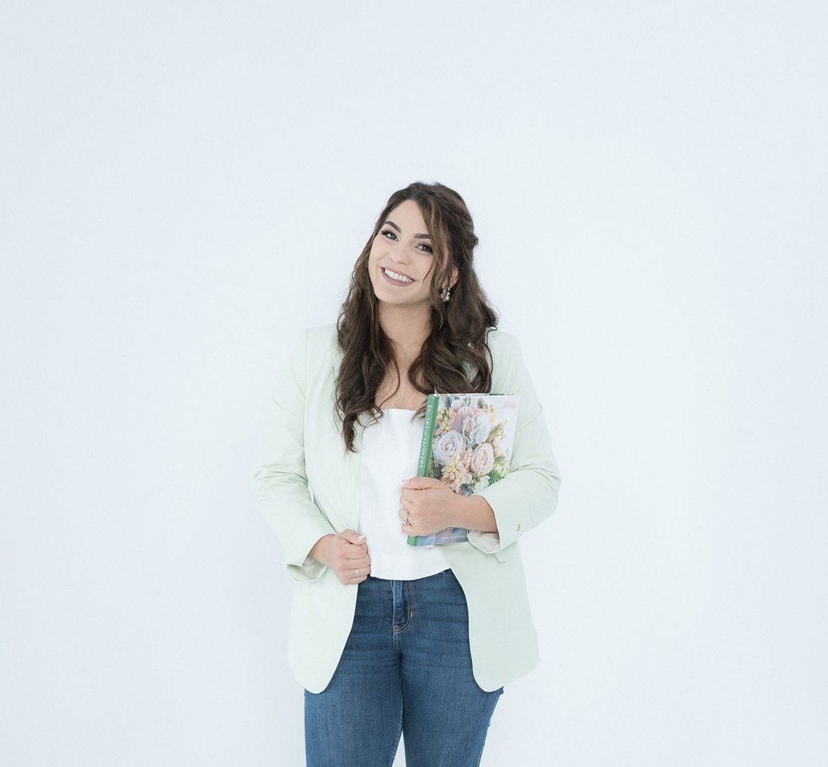 Woman in a light green blazer and jeans smiles, holding a floral book against a white background.