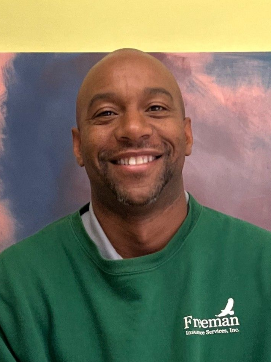 Man smiling, wearing a green sweatshirt with a logo. Against a backdrop of blues, pinks, and yellows.