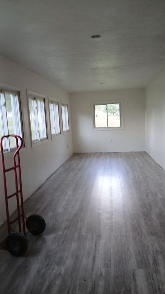 Empty white room with gray wood floor, windows along one wall, and a red hand truck near the left side.