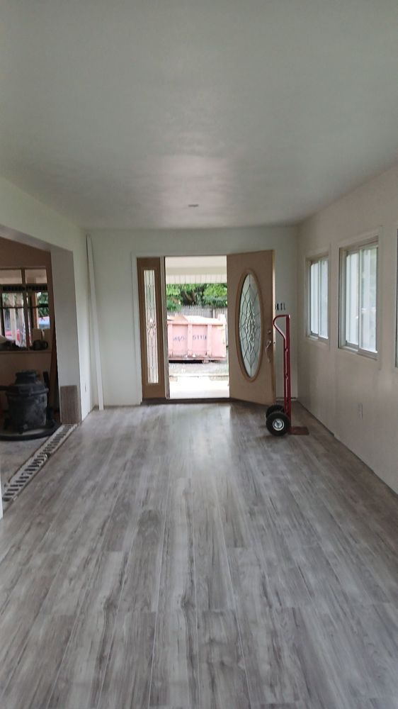 Empty living room with gray wood floors, white walls, front door, and windows letting in daylight