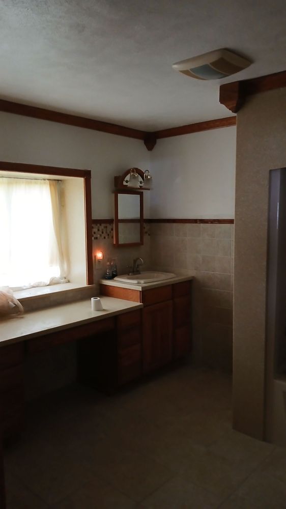 Dim bathroom with vanity, sink, mirror, and sunlit window in a small tiled room.