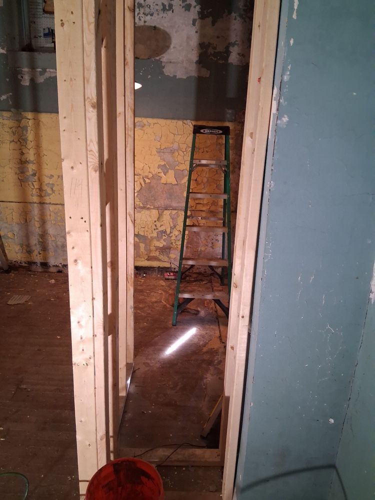 Narrow hallway with exposed studs, worn plaster walls, and a ladder in the back room.