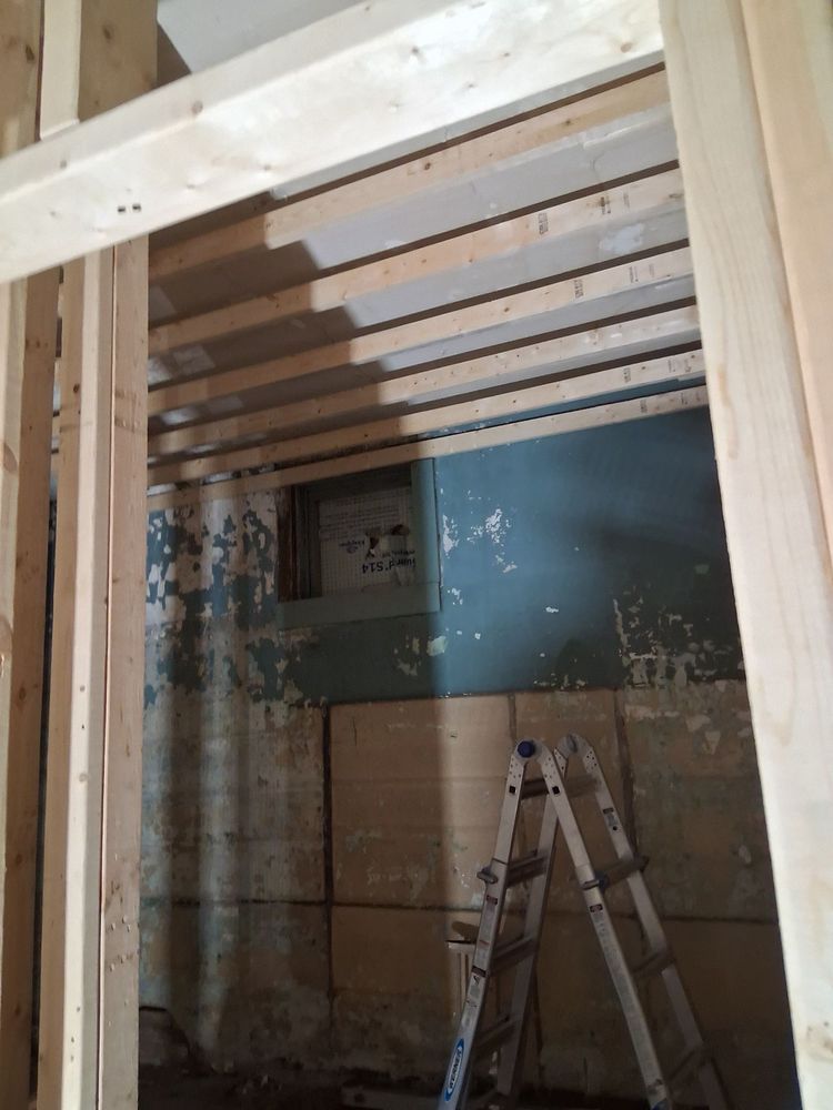 Partially framed interior room with exposed studs, ladder, and stacked plywood against a blue wall