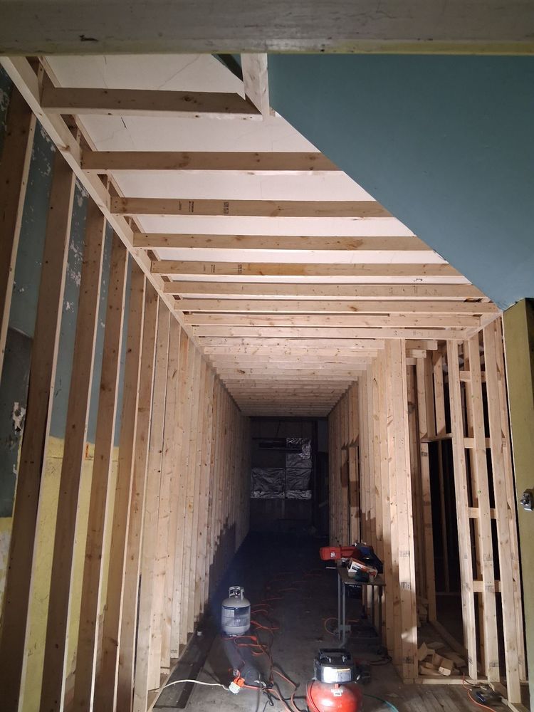Unfinished hallway with exposed wooden framing, ceiling joists, and construction tools on the floor.