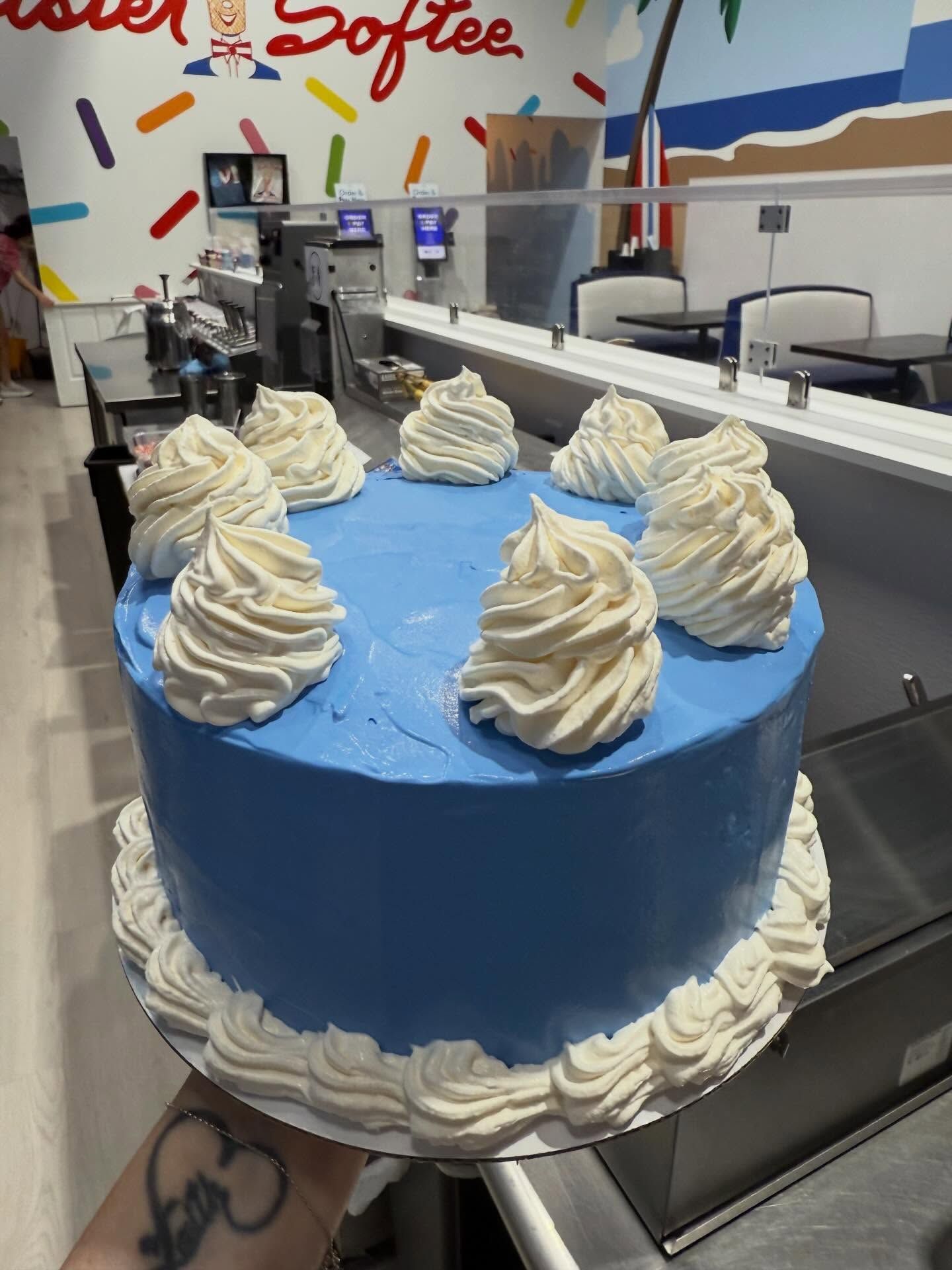 Blue frosted cake with white swirls displayed in a bakery case under colorful sprinkles signage