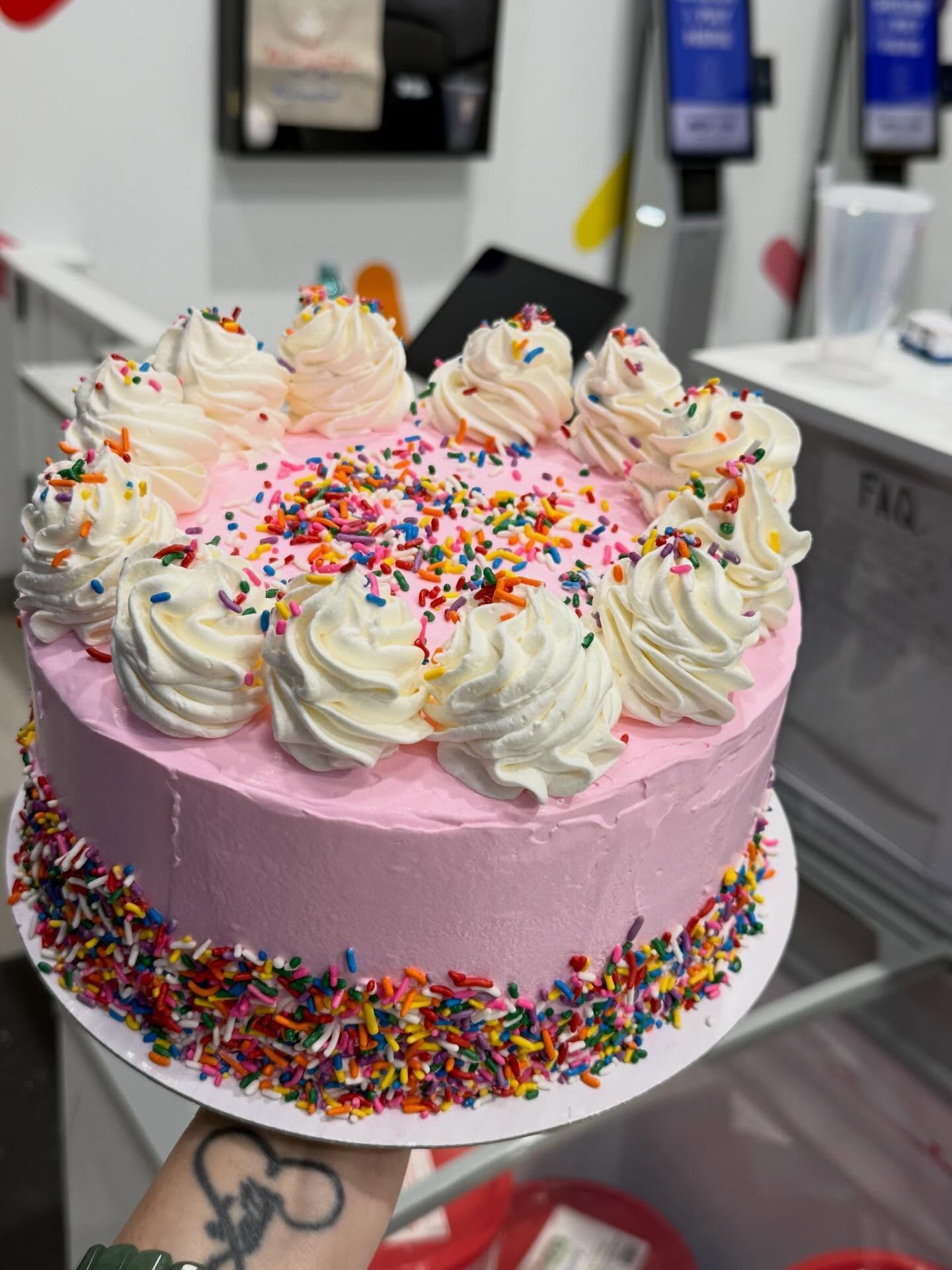 Pink frosted cake topped with whipped cream swirls and colorful sprinkles on a counter