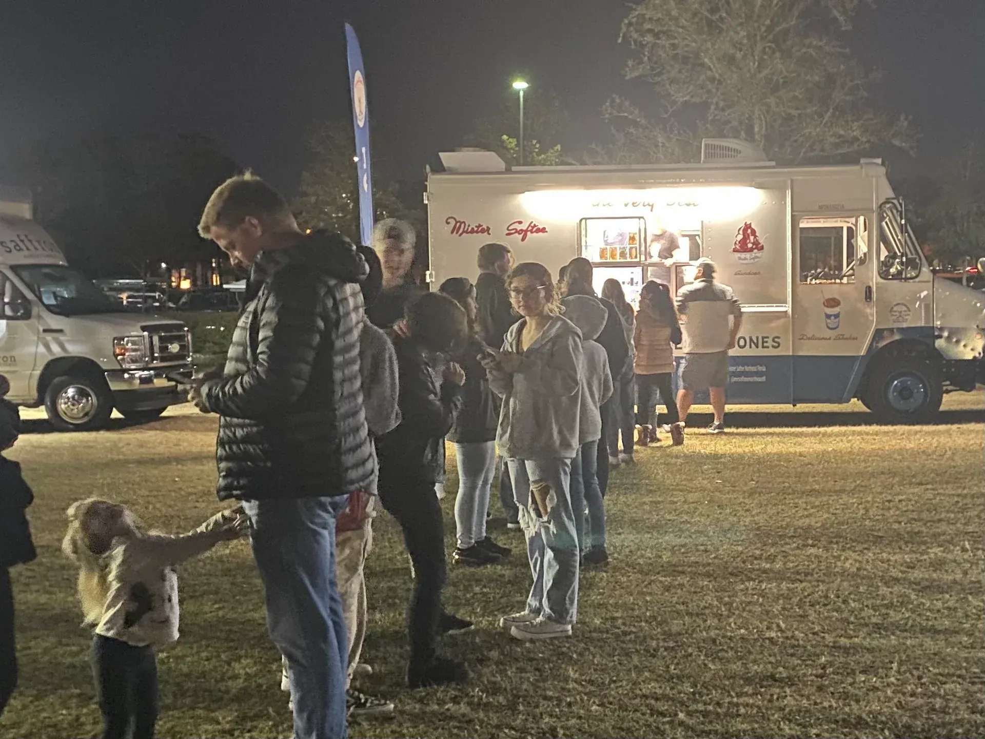 People lined up at a brightly lit food truck at night in a grassy field