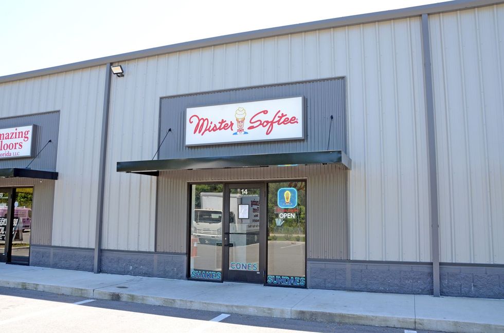Storefront with gray siding and a red-and-white sign above glass double doors.