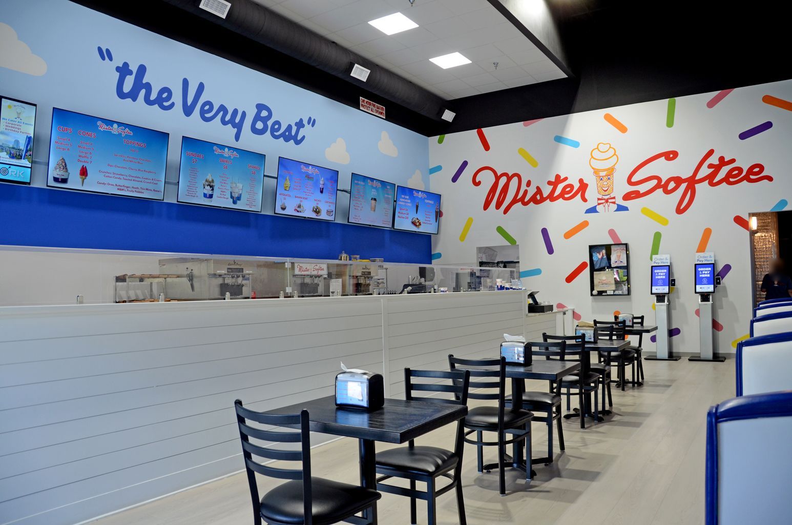 Bright fast-food counter with blue booths, wall mural, menu screens, and empty tables inside a casual restaurant.