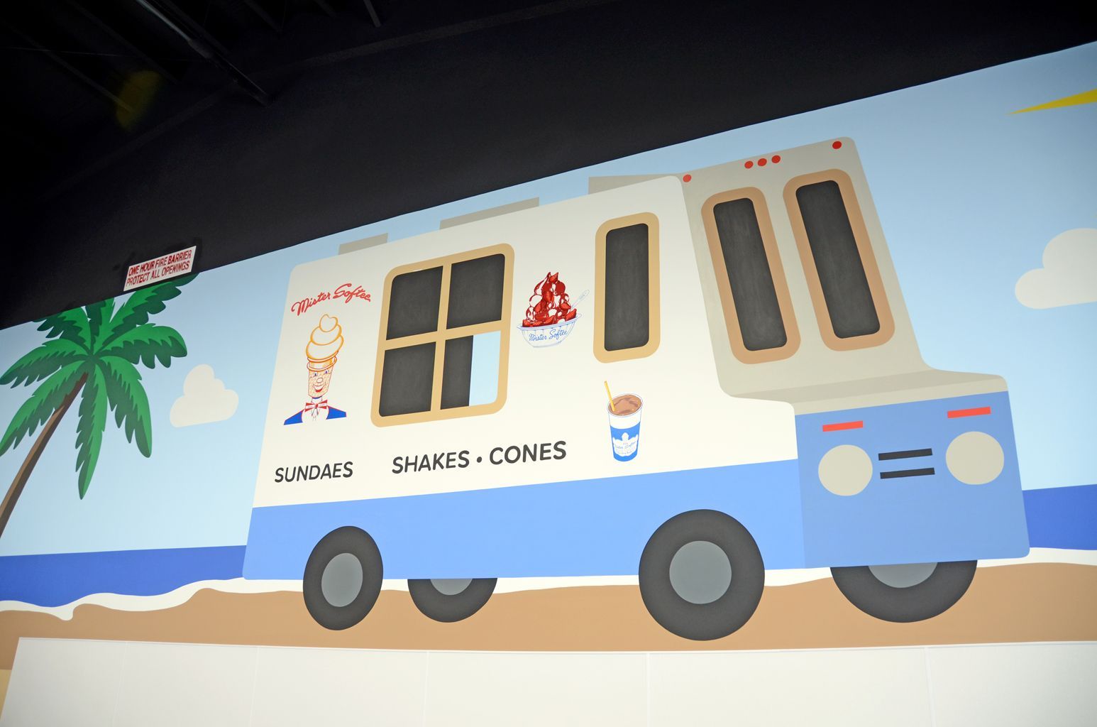 Ice cream truck on a sunny beach, white with blue trim and a palm tree nearby.