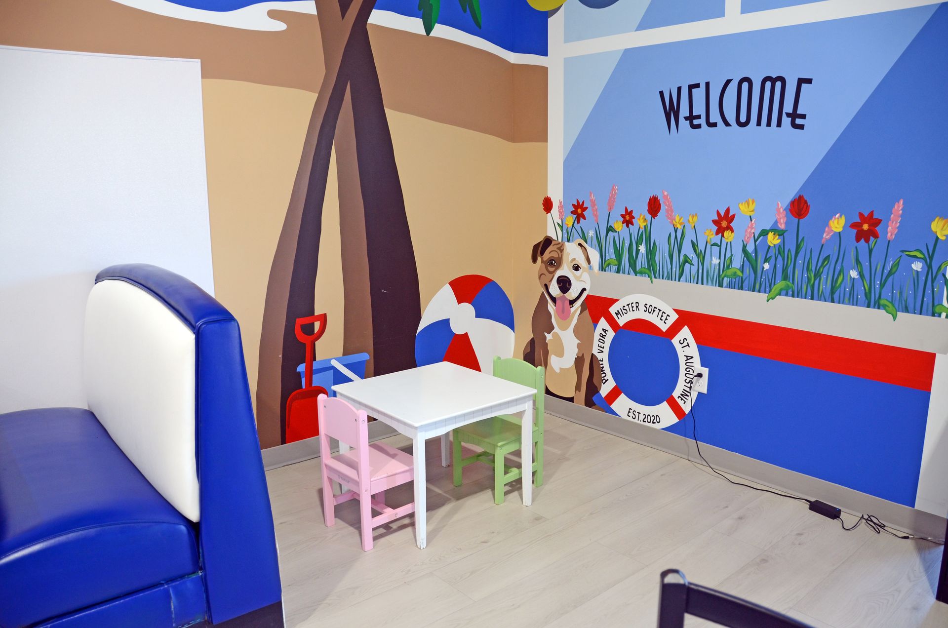Child-sized play area with a blue couch, small table and chairs, and a colorful “WELCOME” wall mural.