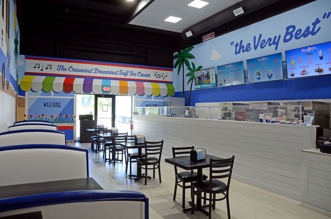 Bright ice cream shop interior with white counter, black tables, and blue walls reading “The Very Best”