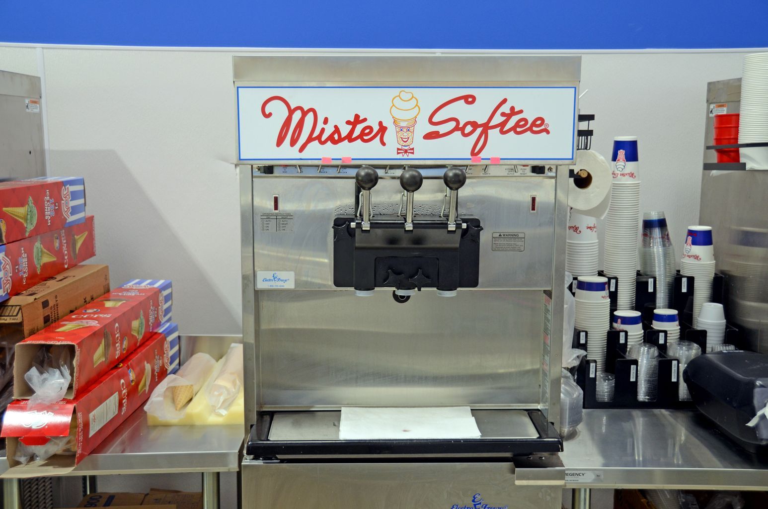 Soft-serve ice cream machine centered on a counter, with stacks of cups and supplies on both sides.