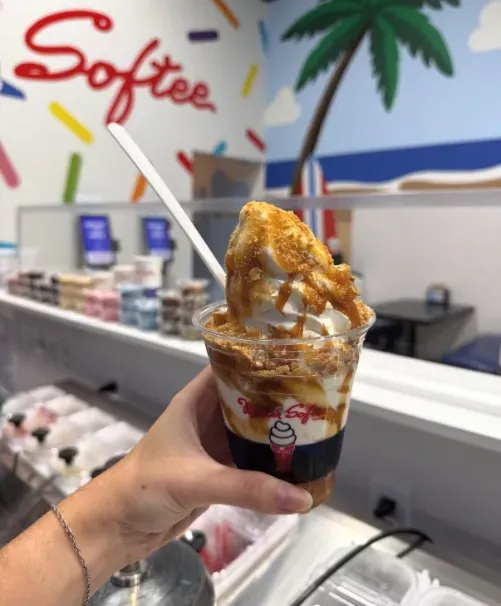 Hand holding a caramel-topped soft serve sundae in a branded cup inside an ice cream shop