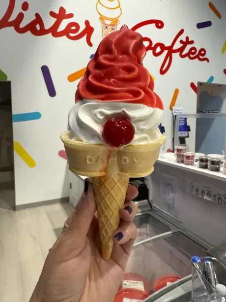 Swirled red-and-white soft-serve cone with a cherry, held inside an ice cream shop