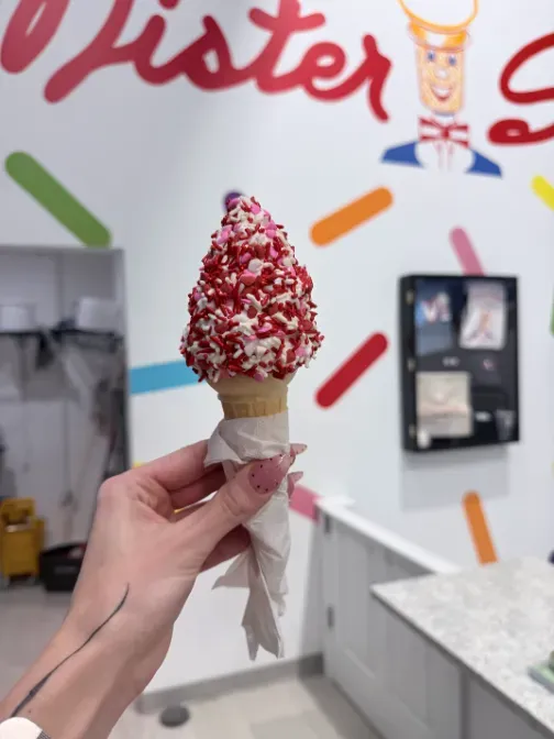 Hand holding a red-and-white sprinkle ice cream cone inside a shop