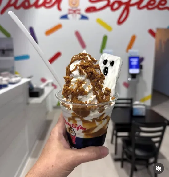 Hand holding a tall dessert cup topped with whipped cream, caramel, and a white chocolate bar in a dessert shop