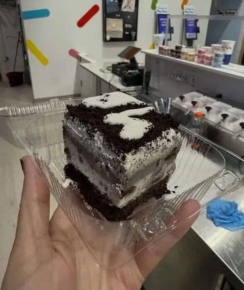 Hand holding a chocolate square dessert in a clear plastic tray at a café counter