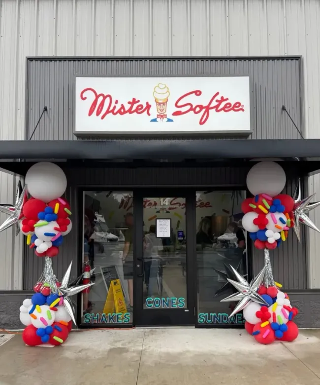Storefront for Mister Softee decorated with colorful balloon bouquets and large white balloons at the entrance.