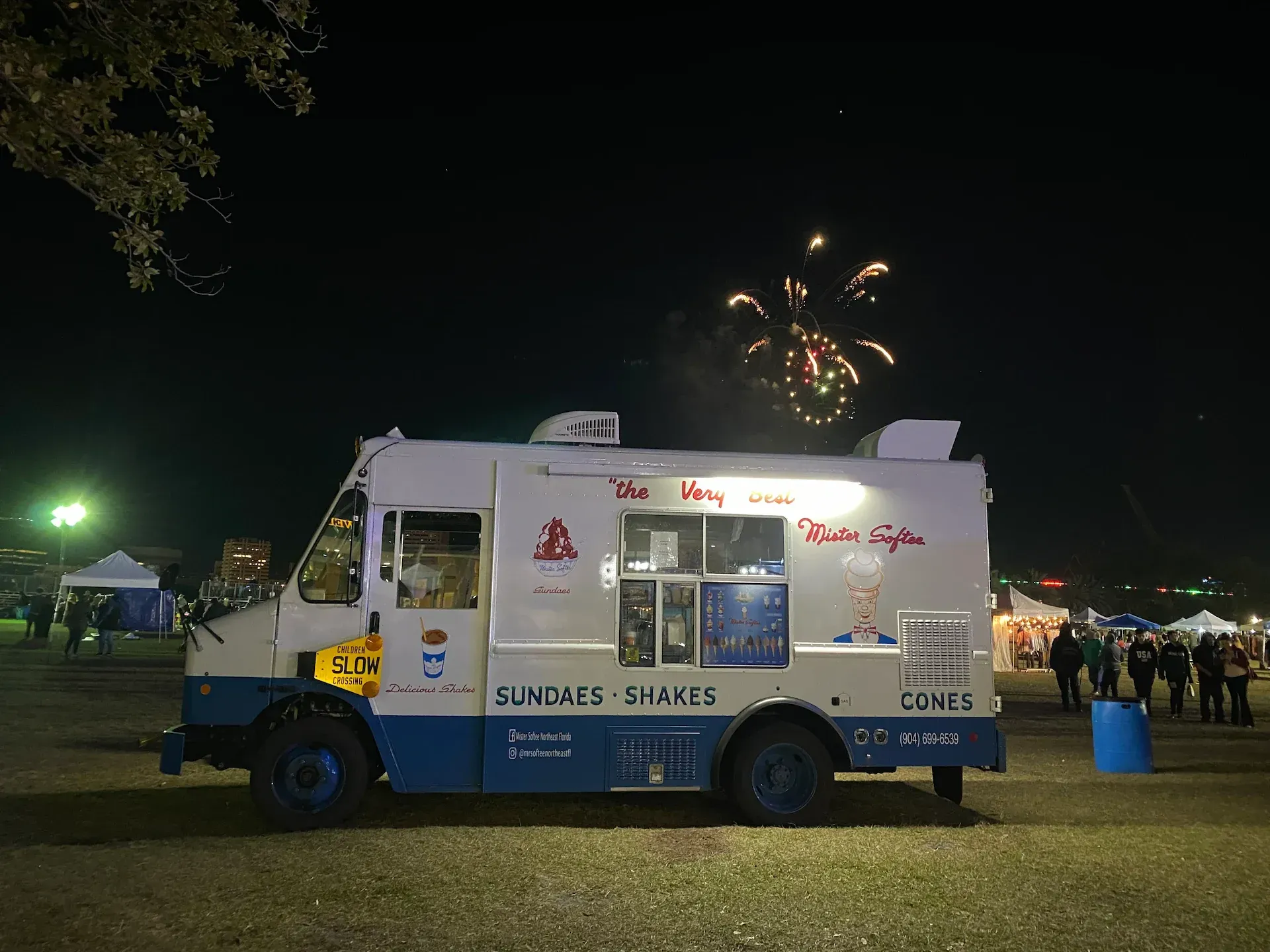 White ice cream truck at a nighttime park with fireworks in the sky.