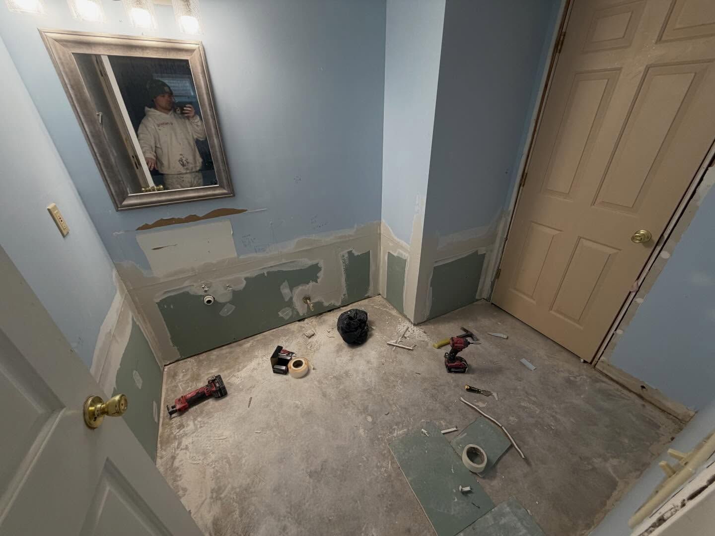 Small blue room under renovation with damaged walls, bare floor, scattered tools, and a closed door.
