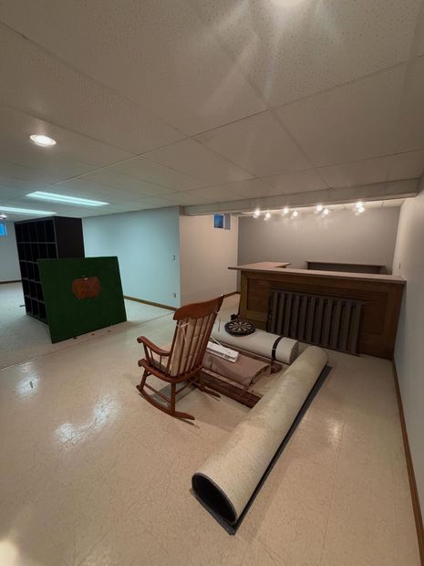 Furniture set in a room under construction, with a rocking chair, bench, rolled rug, and wooden cabinet.