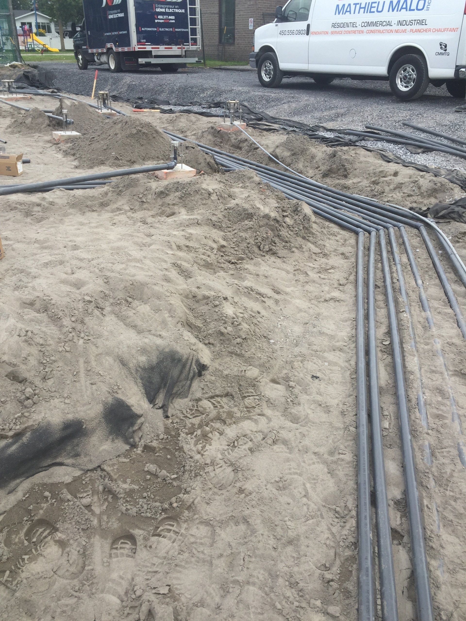 Chantier de construction avec canalisations apparentes dans le sable, camions en arrière-plan.