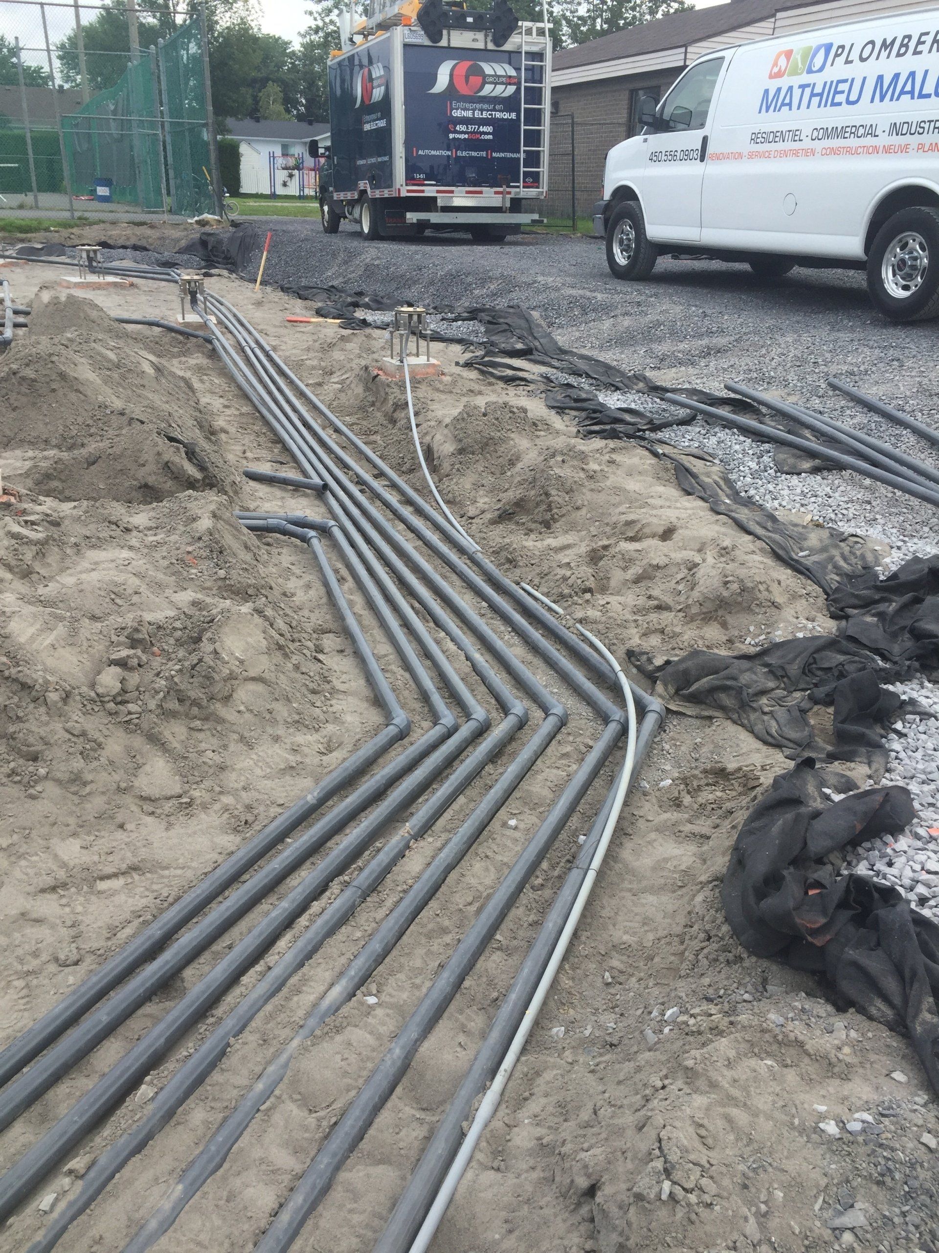 Tuyaux et conduits souterrains sur un chantier de construction, avec un camion utilitaire et une camionnette de plomberie stationnés à proximité.