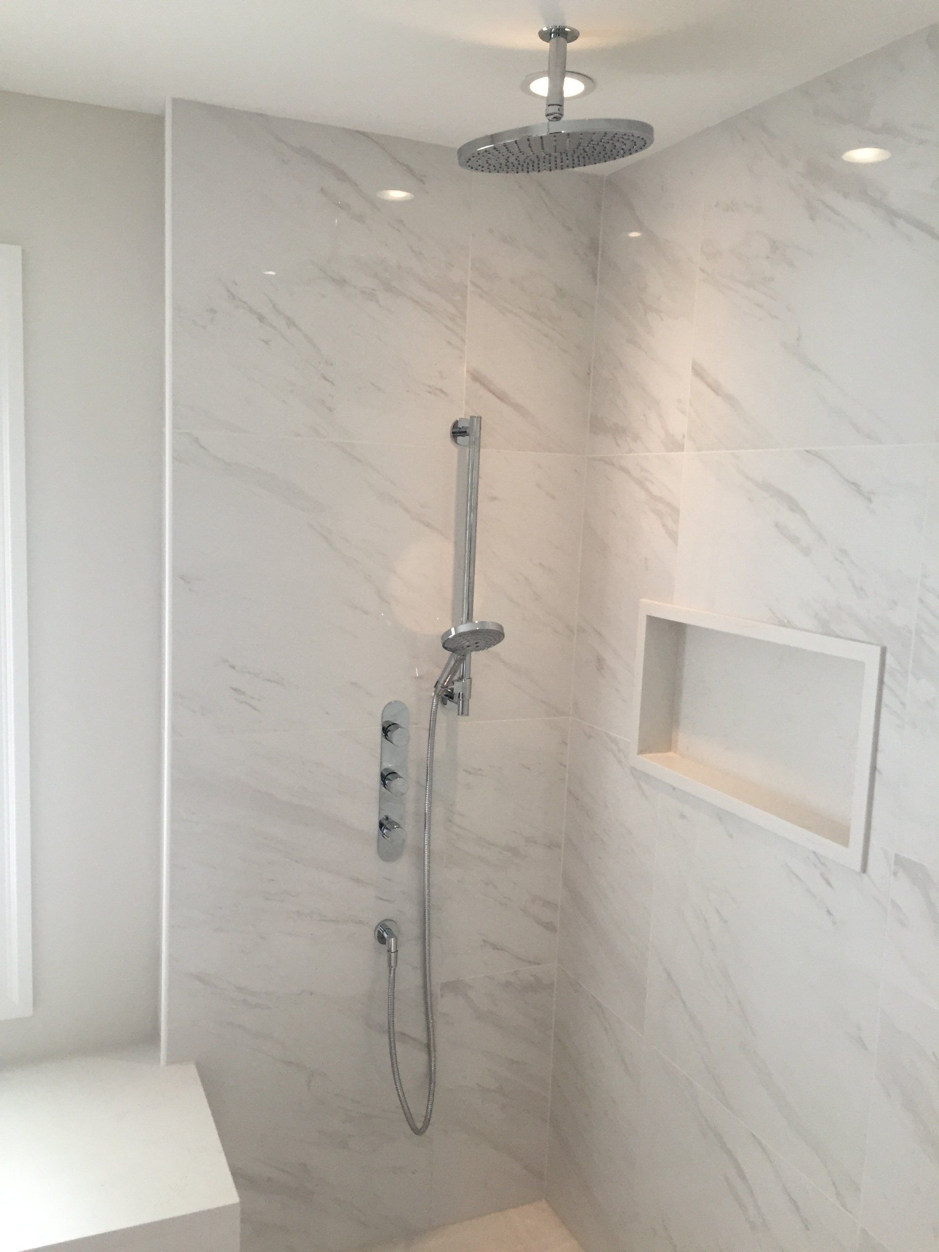 Douche carrelée en marbre blanc avec pommeau de douche effet pluie, douchette et étagère encastrée.