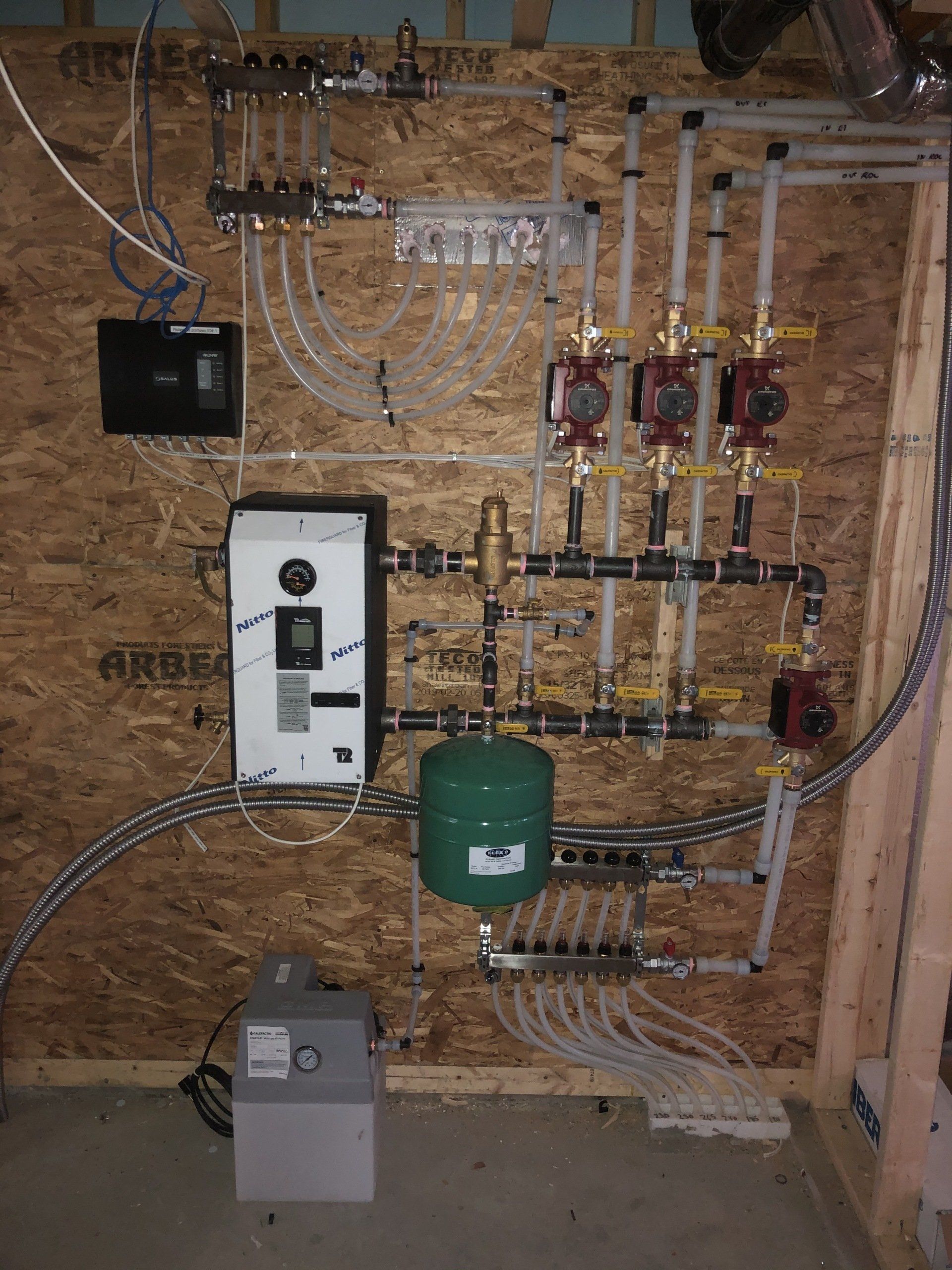 Composants d'un système de chauffage radiant montés sur un mur en bois, comprenant des pompes, des collecteurs et un réservoir sous pression.