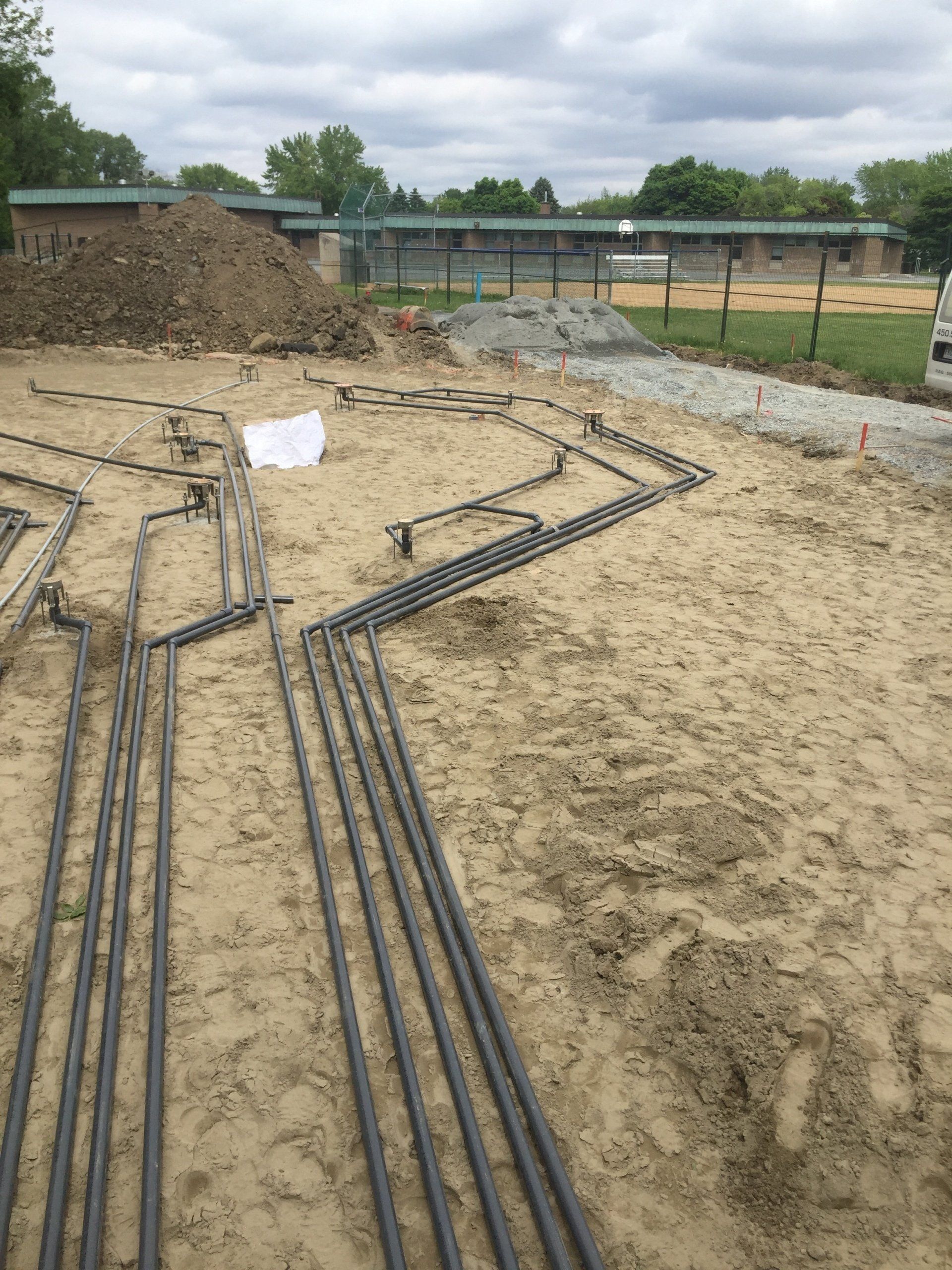 Chantier : tuyaux métalliques posés dans des tranchées, monticule de terre, bâtiment en arrière-plan, ciel nuageux.