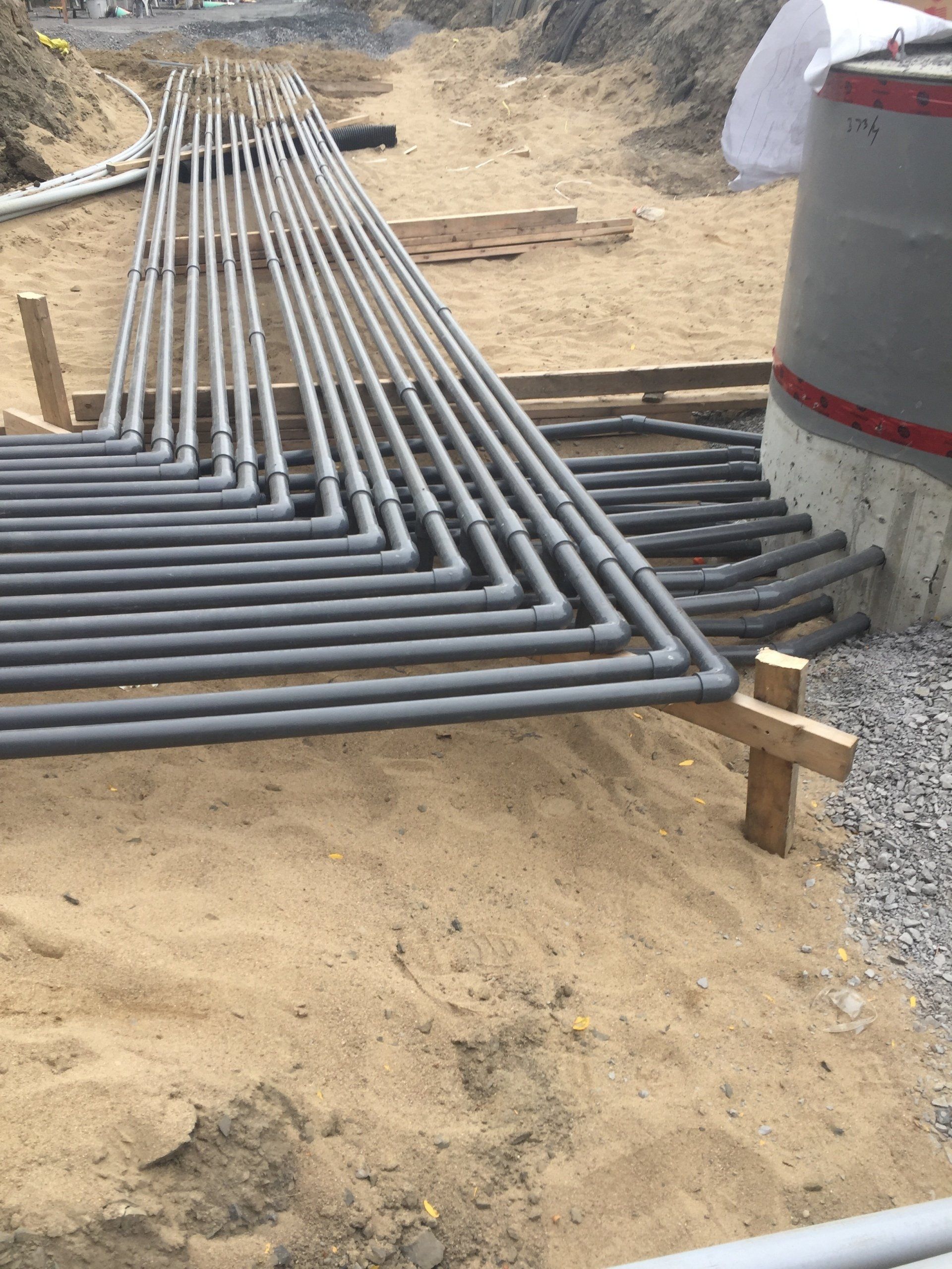 Des tuyaux gris, coudés à angle droit, reposent dans une tranchée sablonneuse près d'une structure en béton, soutenue par des piquets en bois.