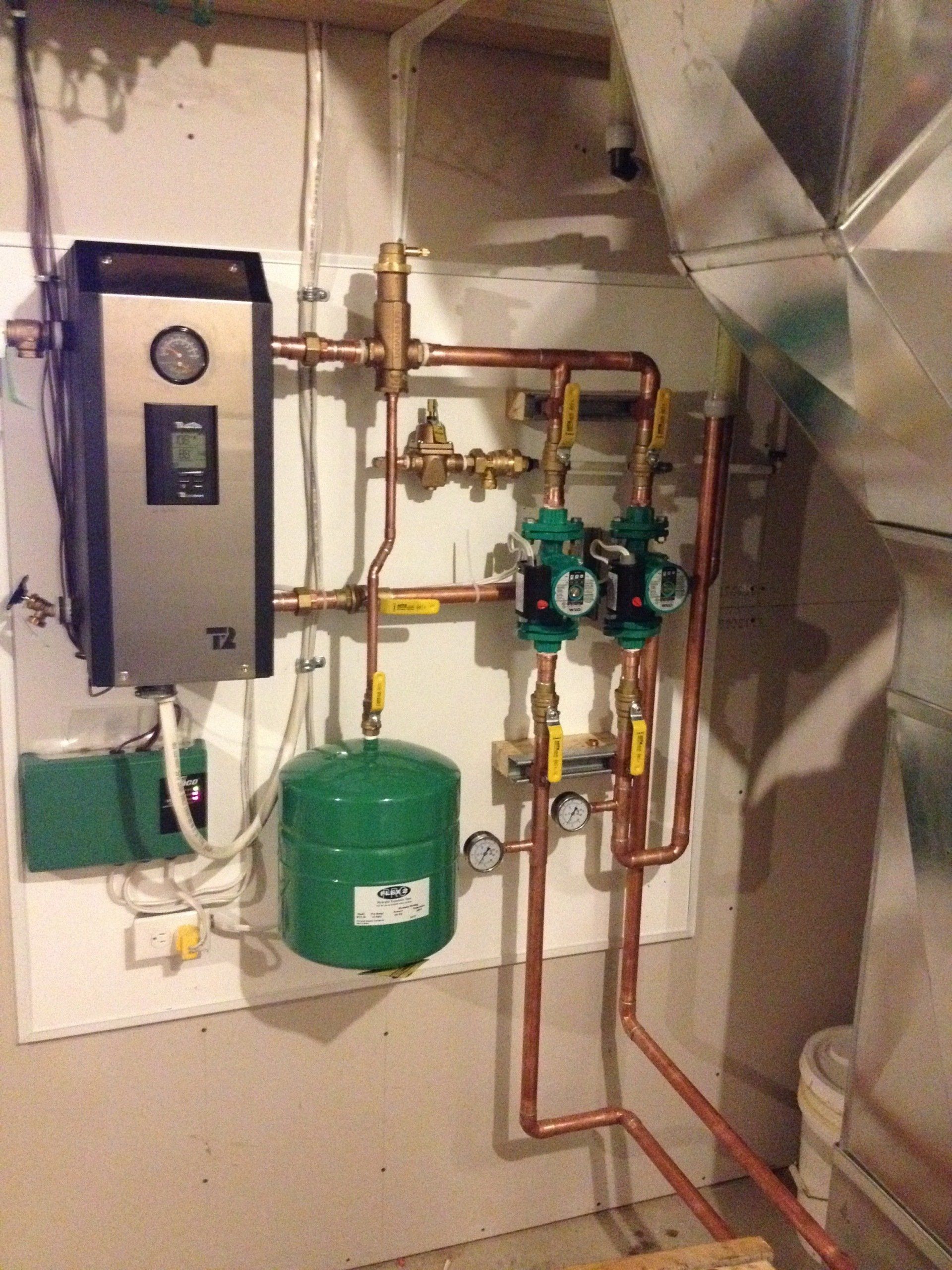 Système de chauffage avec panneau de commande noir, tuyaux en cuivre, pompes vertes et vase d'expansion vert.