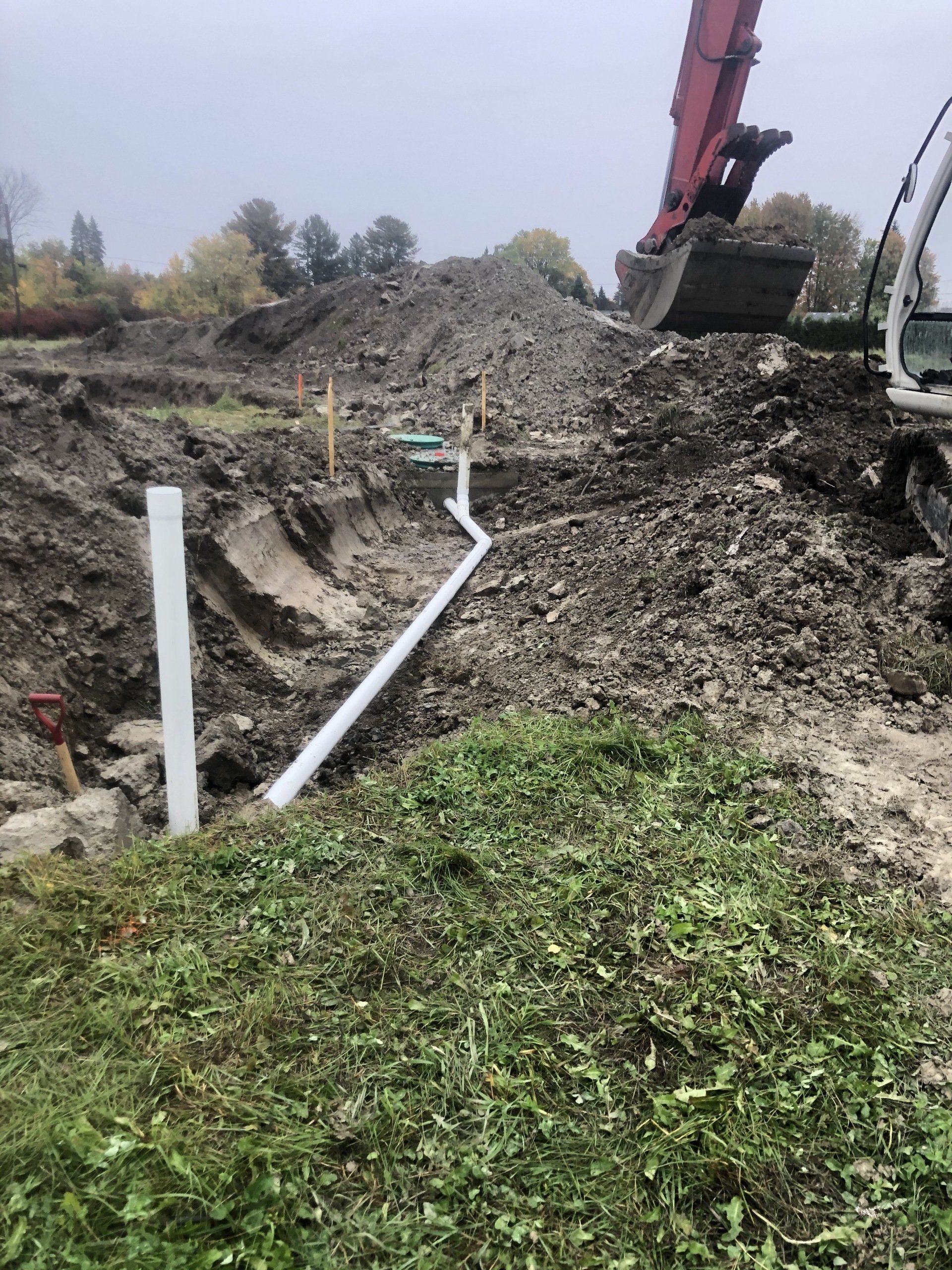 Chantier de construction avec un tuyau blanc posé dans une tranchée, une excavatrice et des tas de terre.