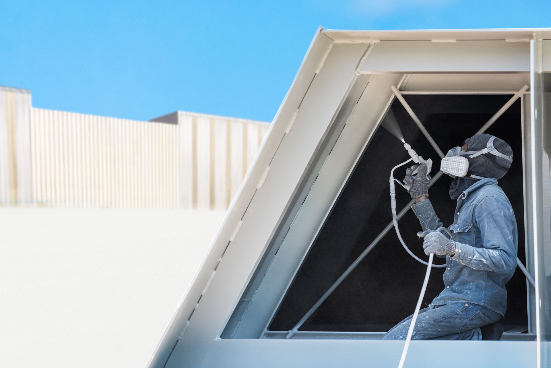 Person in protective gear sprays paint on an architectural structure against a blue sky.