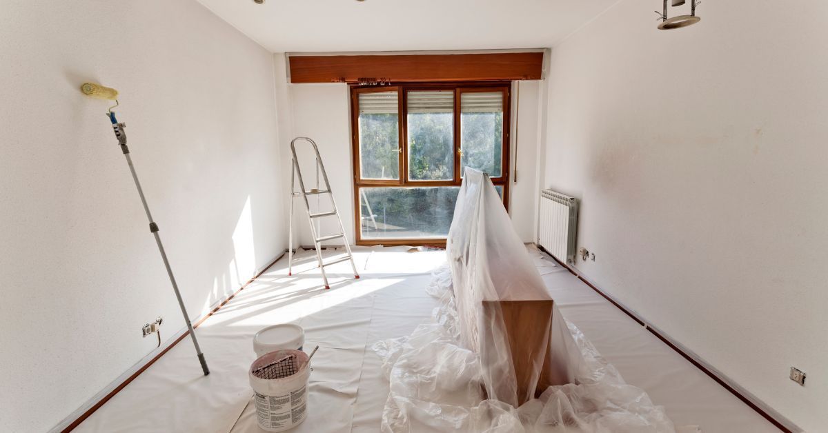 Room being painted with roller, ladder, window, and covered furniture. White walls, wood trim.