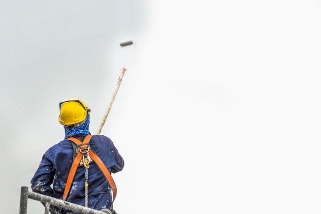 Painter in safety harness using roller to paint a white surface.