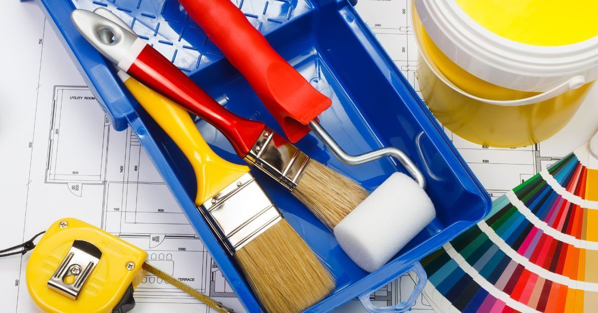 Paint brushes, roller, paint can, and color swatches in a blue tray on a blueprint. Yellow and red accents.