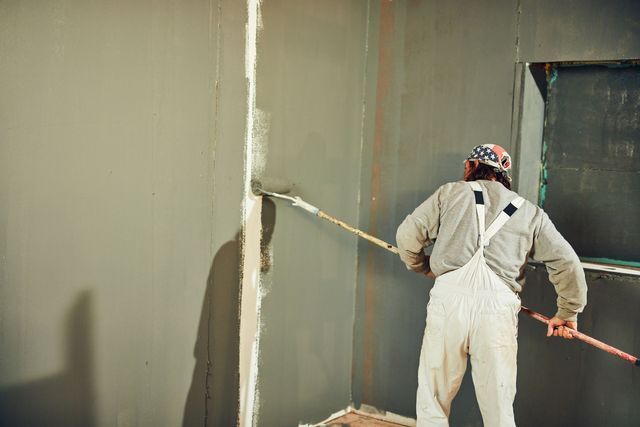 Painter in overalls rolls gray paint onto a wall. Corner of room, partially painted.