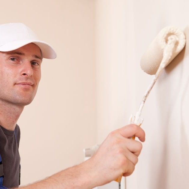 Man in white cap painting a wall with a roller. Beige wall and paint.